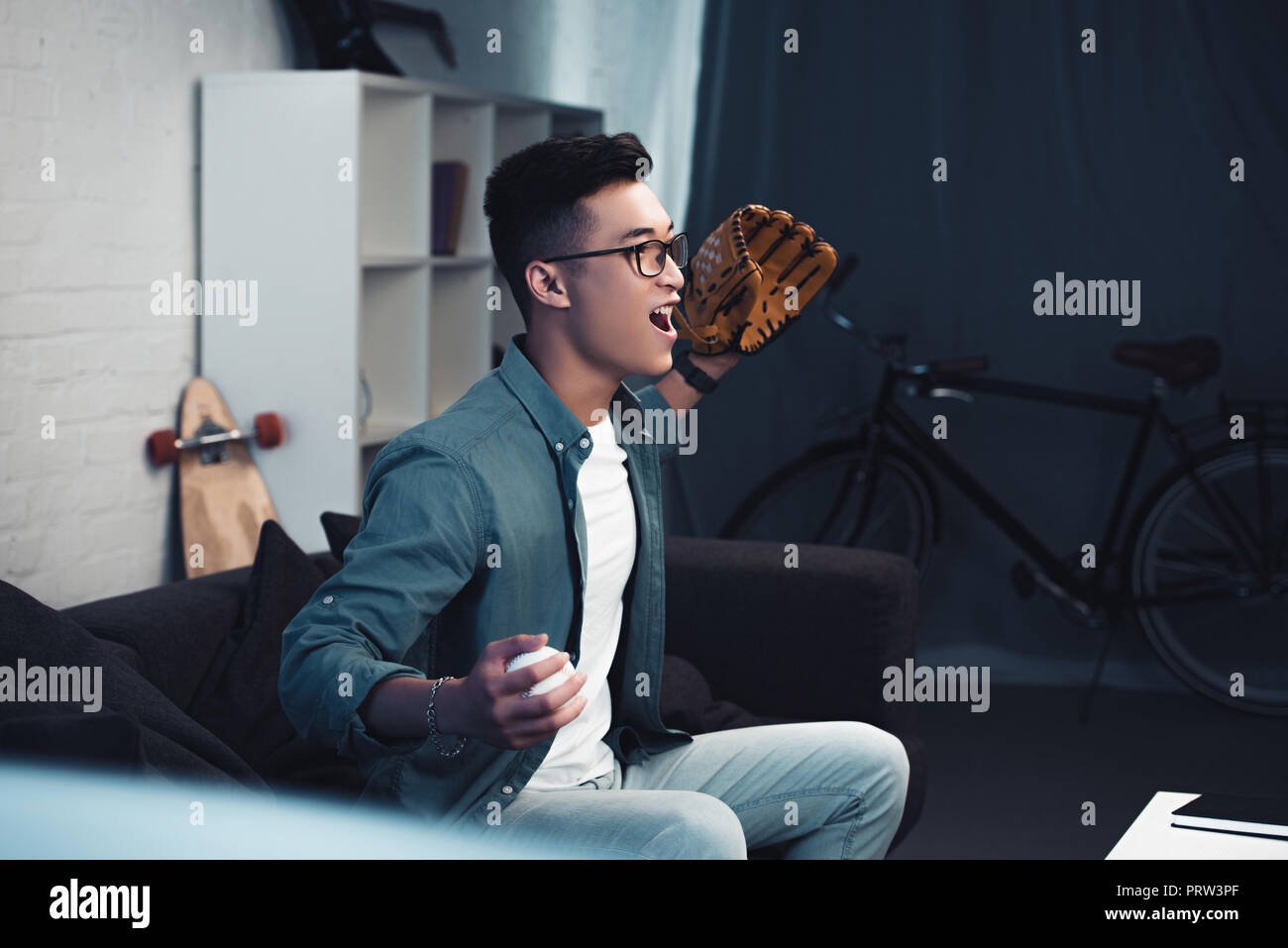 emotional young asian man with baseball glove and ball sitting on couch and watching sport match at home Stock Photo