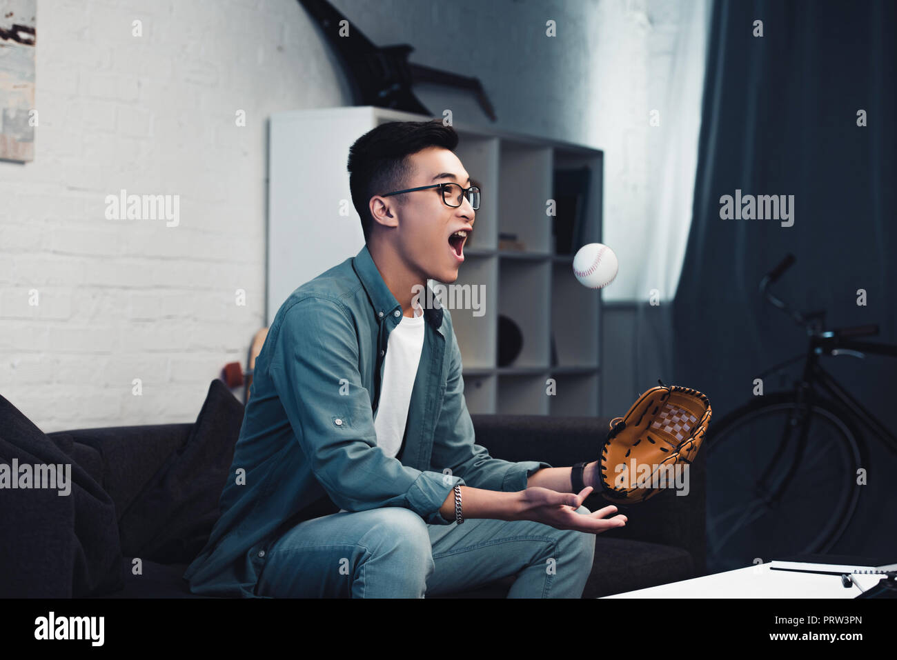 emotional young asian man with baseball glove and ball sitting on couch and watching sport match at home Stock Photo
