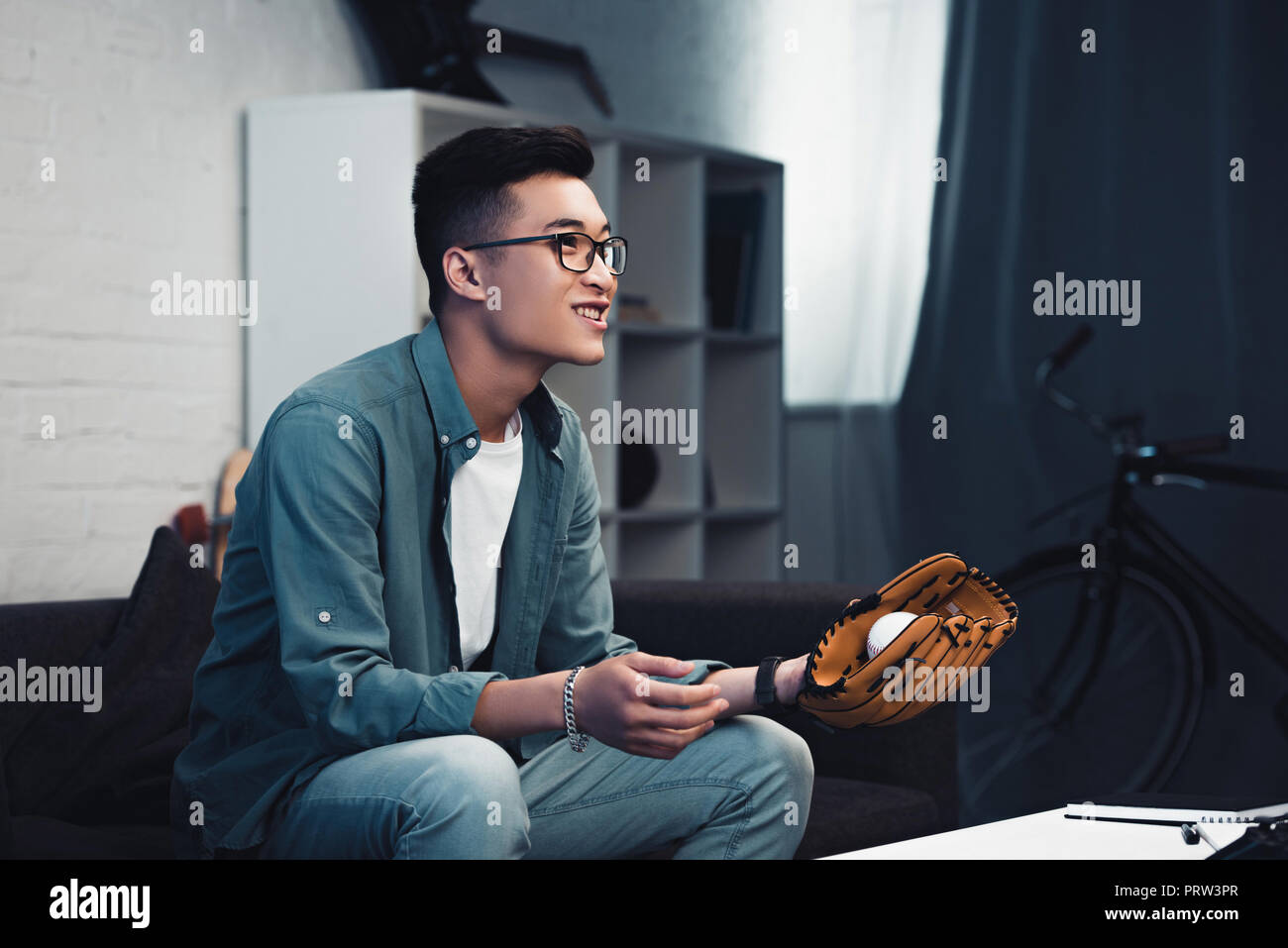 smiling young asian man with baseball glove and ball sitting on couch and watching sport match at home Stock Photo