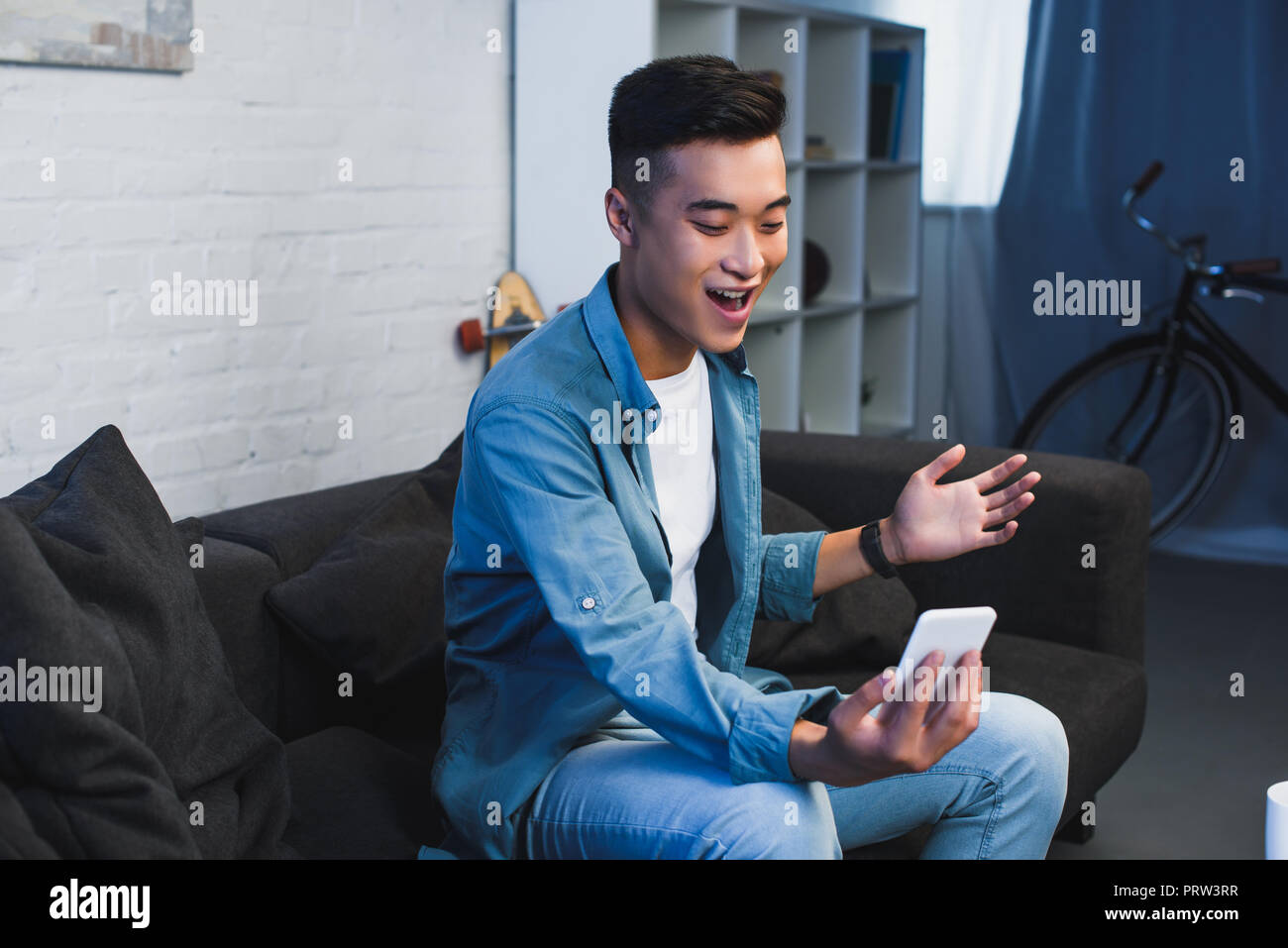 smiling young asian man sitting on couch and using smartphone at home Stock Photo
