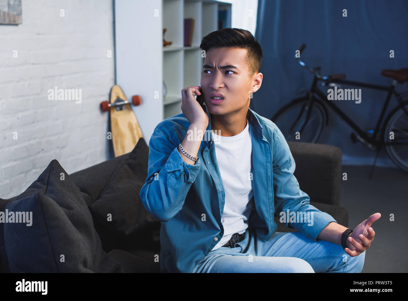 upset young man sitting on couch and talking by smartphone at home Stock Photo