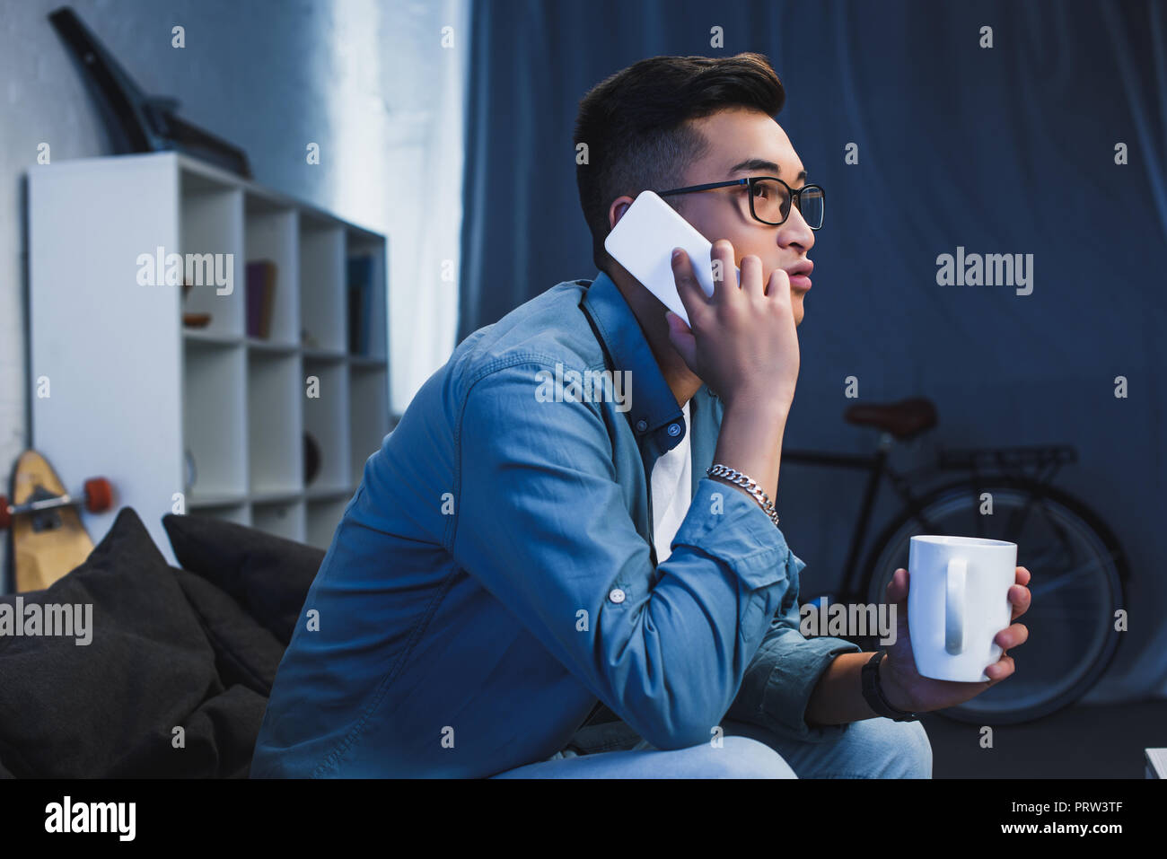 side view of young asian man in eyeglasses holdign cup and talking by smartphone Stock Photo