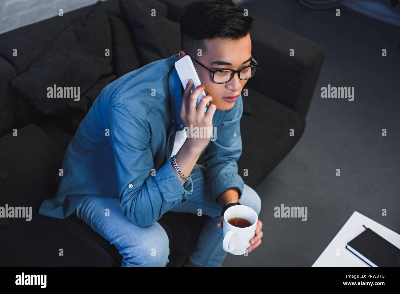 high angle view of young asian man in eyeglasses holdign cup of tea and talking by smartphone Stock Photo