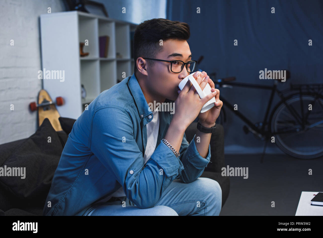 young asian man in eyeglasses sitting on couch and drinking from cup Stock Photo