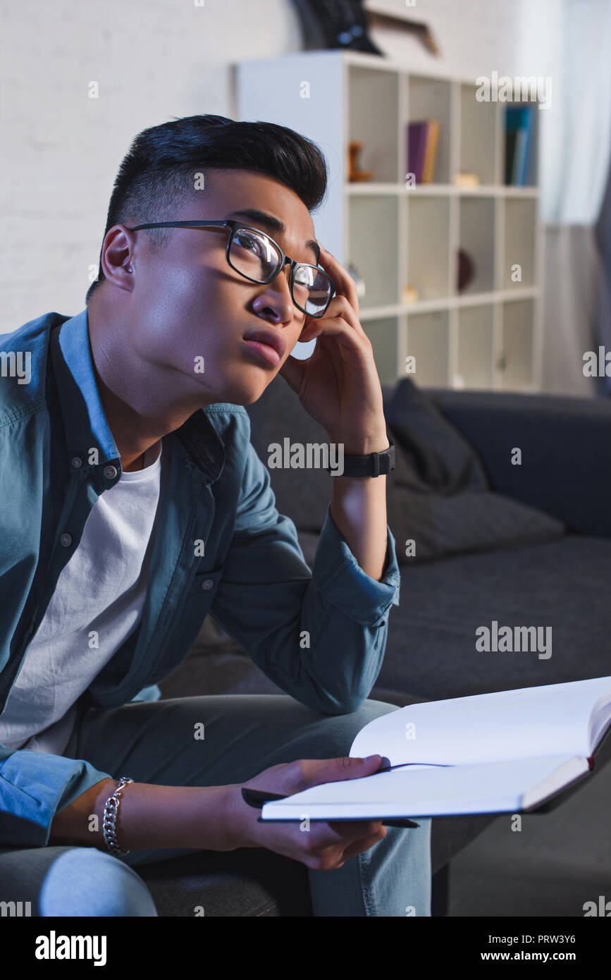 pensive young asian man in eyeglasses holding notebook and looking away at home Stock Photo
