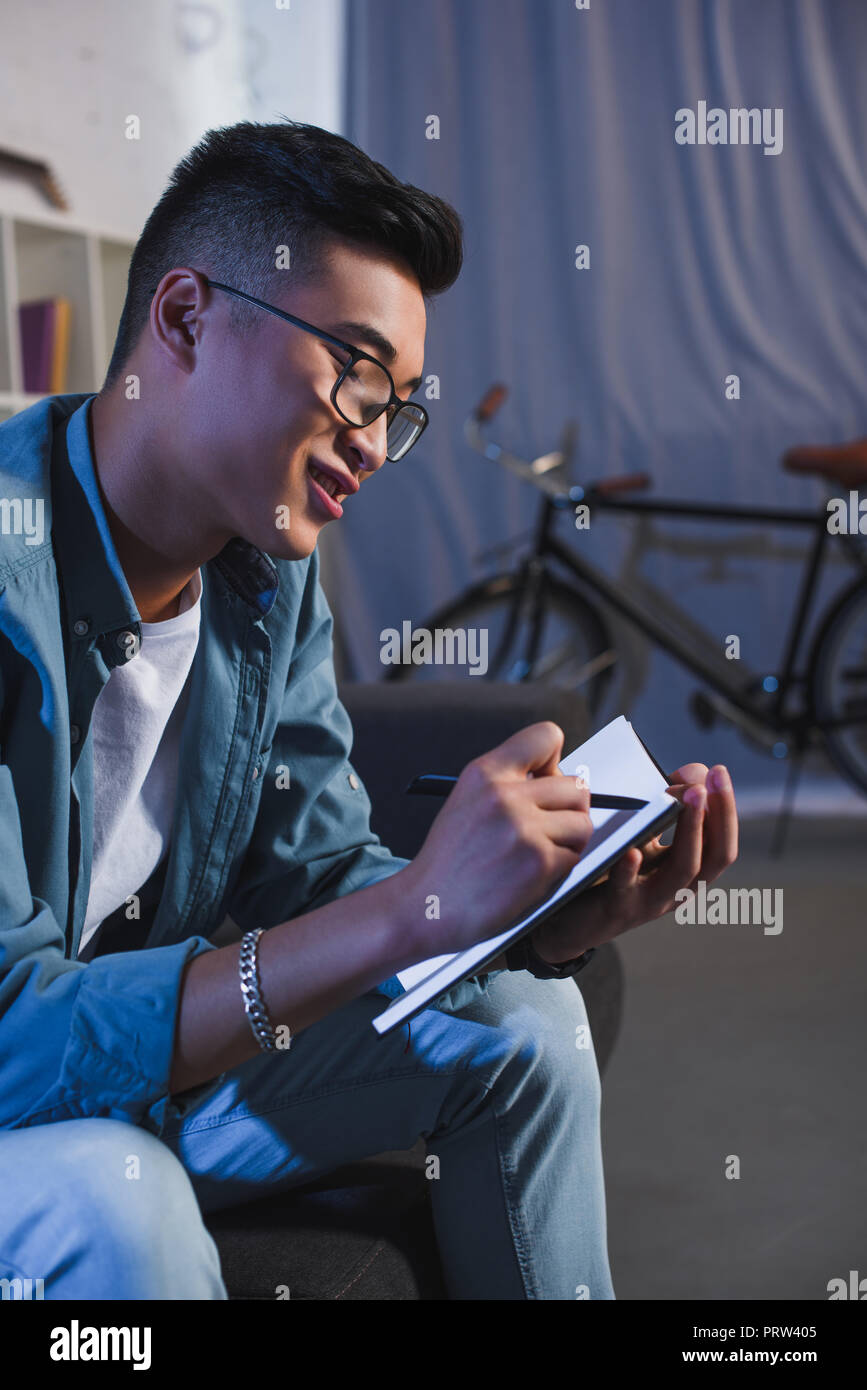 smiling young asian man in eyeglasses writing in notebook at home Stock Photo
