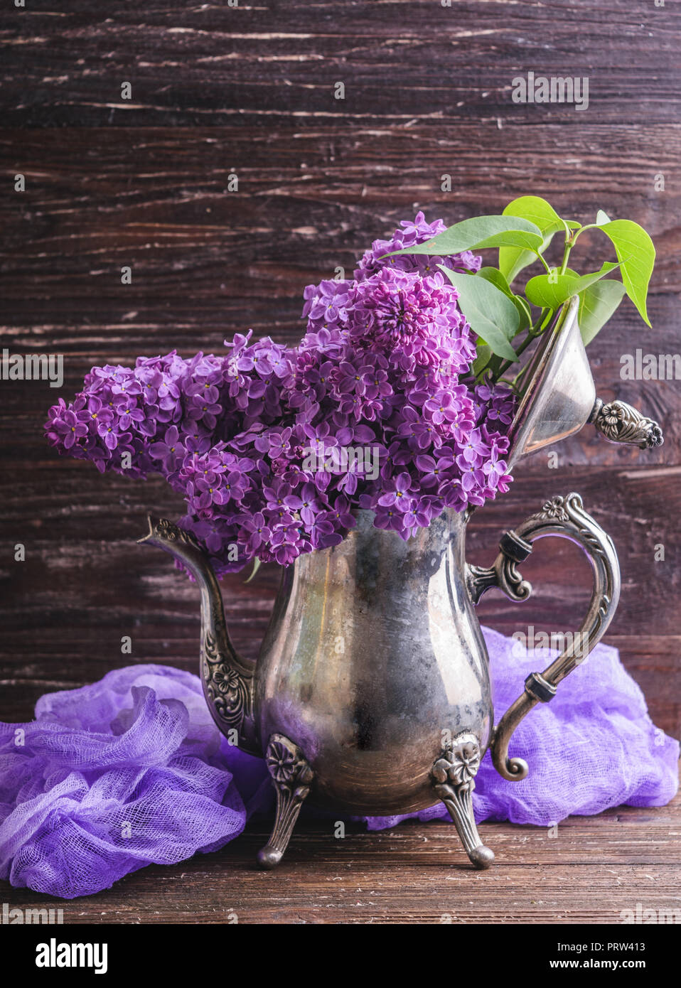 Bouquet of purple lilac flowers in vintage decorative teapot with purple dyed gauze fabric. Dark brown wooden background. Spring romantic flowers deco Stock Photo