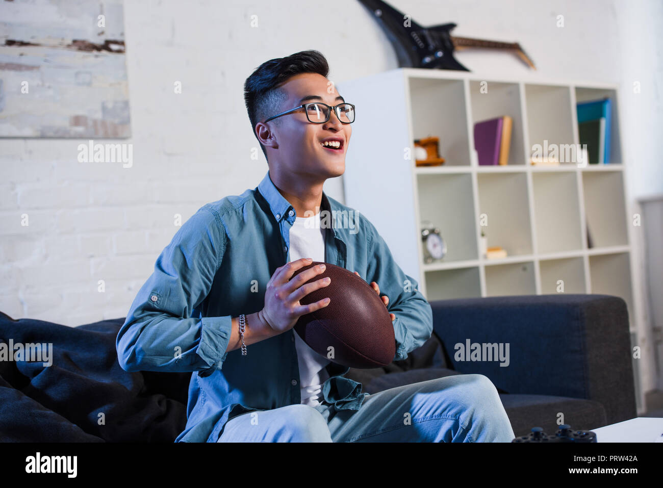 happy young asian man in eyeglasses holding rugby ball and watching sport match at home Stock Photo