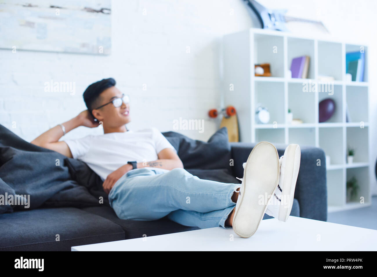 selective focus of handsome asian man sitting on sofa with legs on table at home, looking away Stock Photo