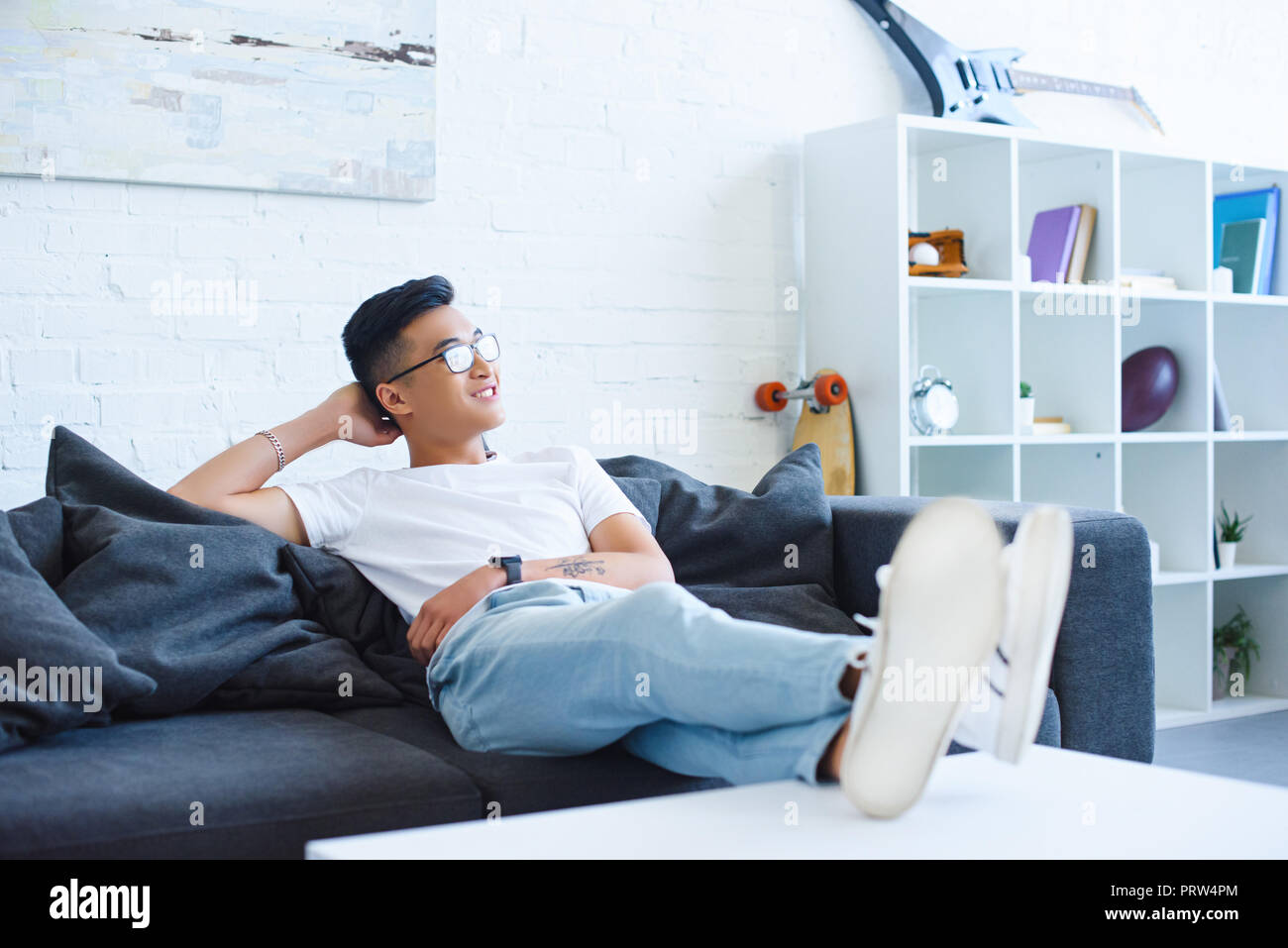 smiling handsome asian man sitting on sofa with legs on table at home, looking away Stock Photo