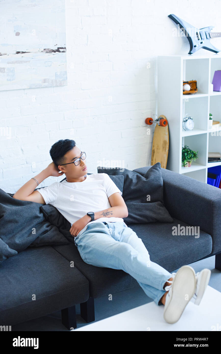 high angle view of pensive handsome asian man sitting on sofa with legs on table at home, looking away Stock Photo