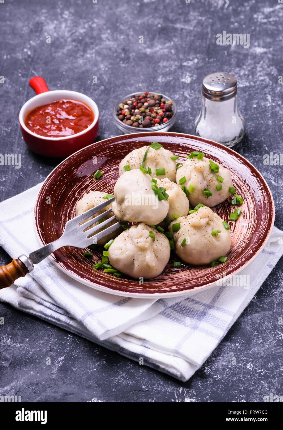Georgian dumplings Khinkali with meat, green onion and tomato sauce. Served on brown ceramic plate with bowl of sauce, pepper mix and salt. Dark concr Stock Photo