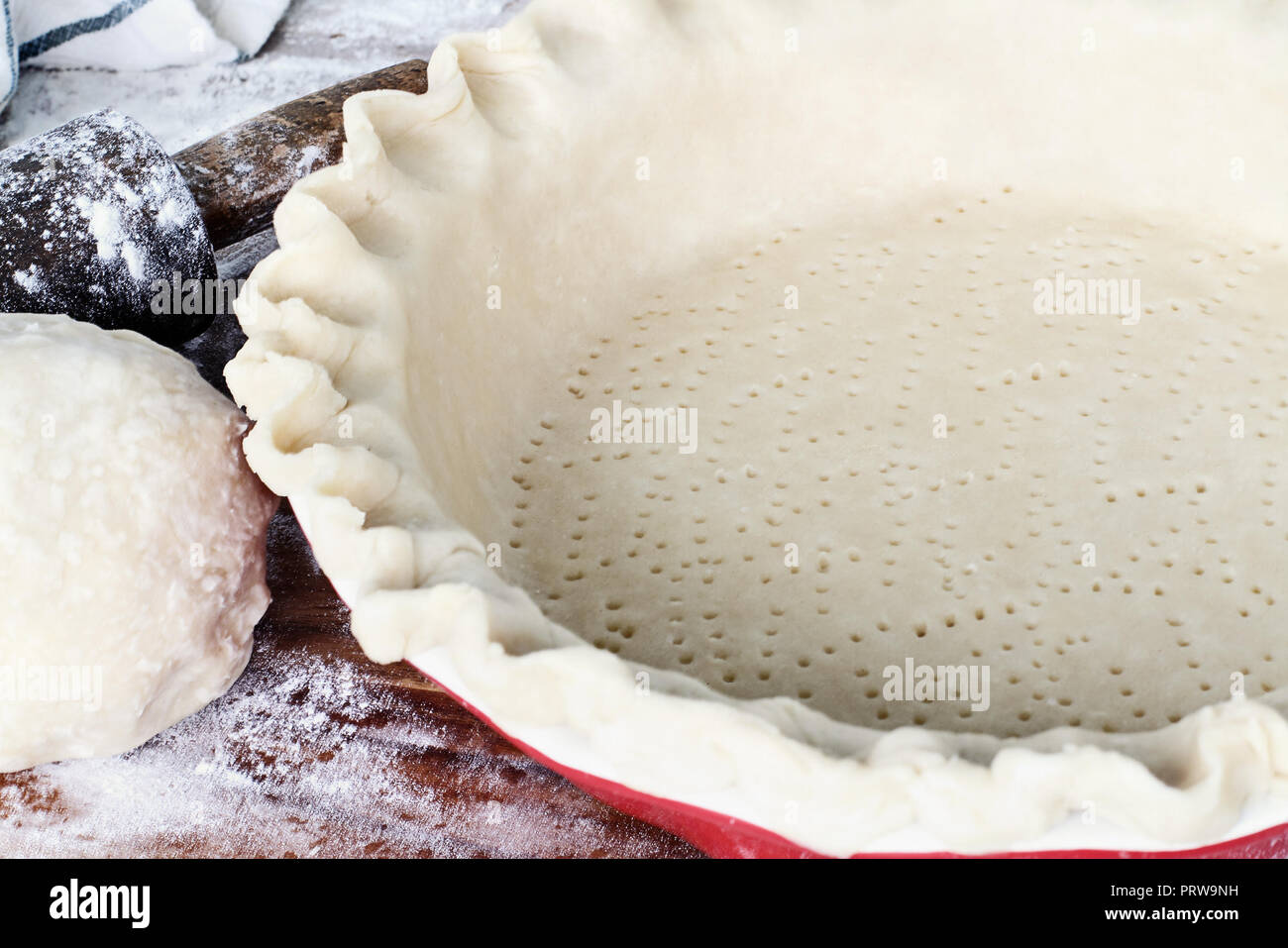 Homemade butter pie crust in pie plate with fluted pinched edge, rolling pin and extra ball of dough over floured rustic wooden background. Crust has  Stock Photo