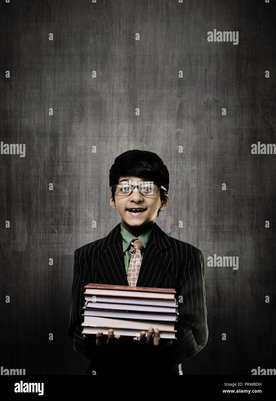 Cute Intelligent Little Boy Holding Books And Wearing Glasses, Smiling While Standing Before A Chalkboard, Stock Photo