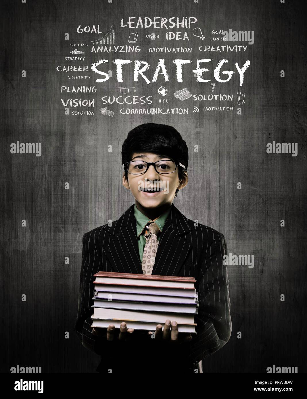 Cute Intelligent Little Boy Holding Books And Wearing Glasses, Smiling While Standing Before A Chalkboard,  Strategy written on board Stock Photo