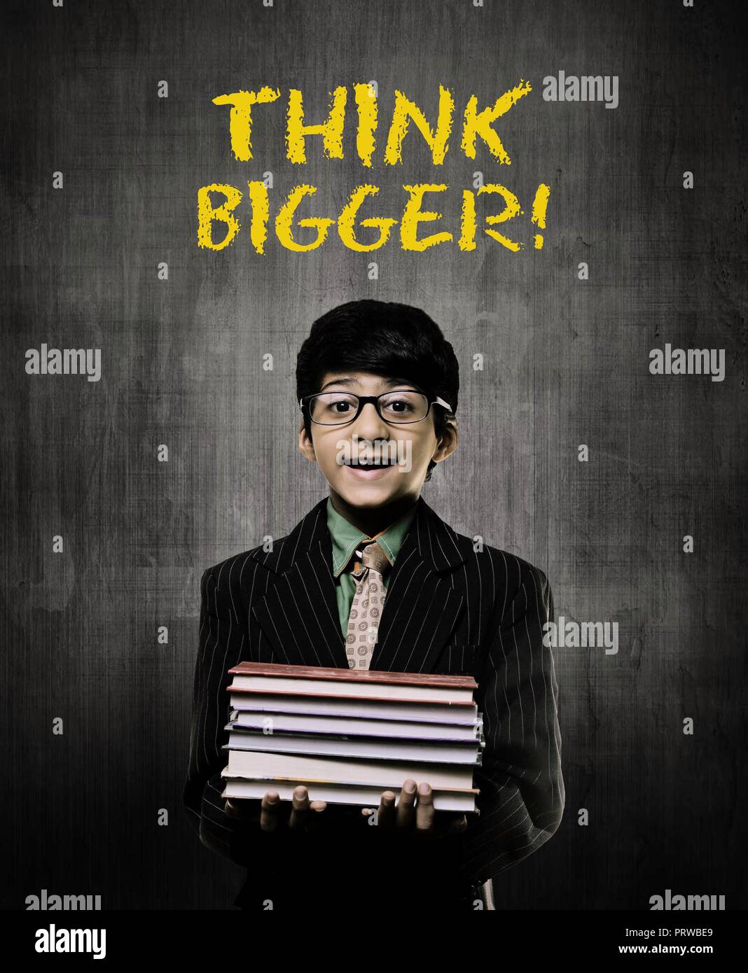 Cute Intelligent Little Boy Holding Books And Wearing Glasses, Smiling While Standing Before A Chalkboard, Think Big Is Written On Board In Yellow Col Stock Photo