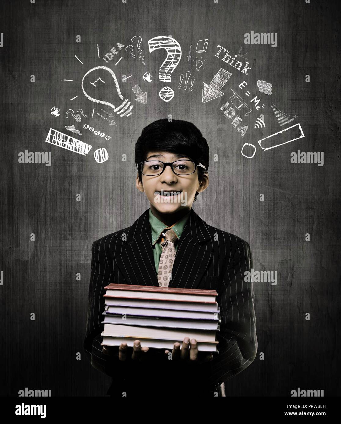 Cute Intelligent Little Boy Holding Books And Wearing Glasses, Smiling While Standing Before A Chalkboard, Thinking Process Elements Drawn Above His H Stock Photo