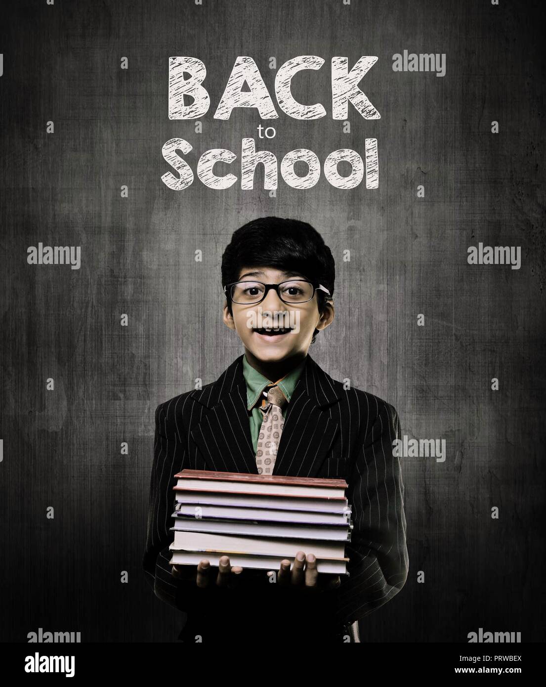 Cute Intelligent Little Boy Holding Books And Wearing Glasses, Smiling While Standing Before A Chalkboard, Back To School Written On Board Stock Photo
