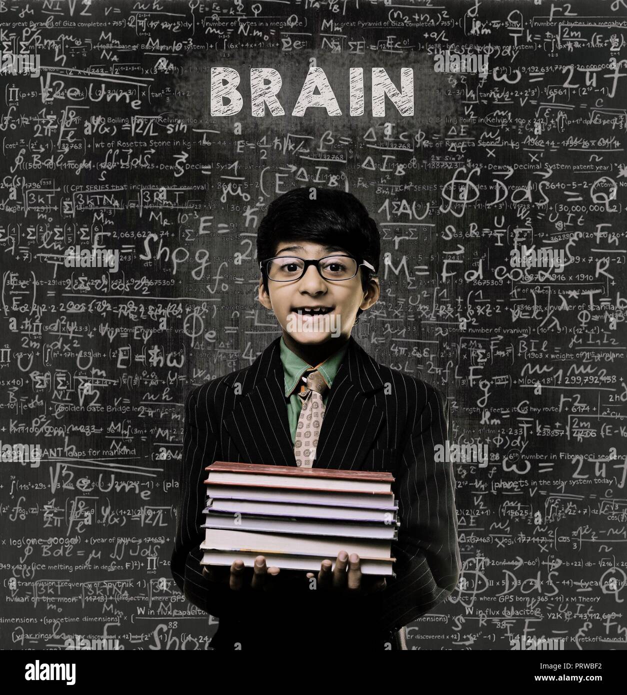 Cute Intelligent Little Boy Holding Book And Wearing Glasses Standing Before A Chalkboard, Chemical Formulas Are Written On Board With The Chalkboard  Stock Photo