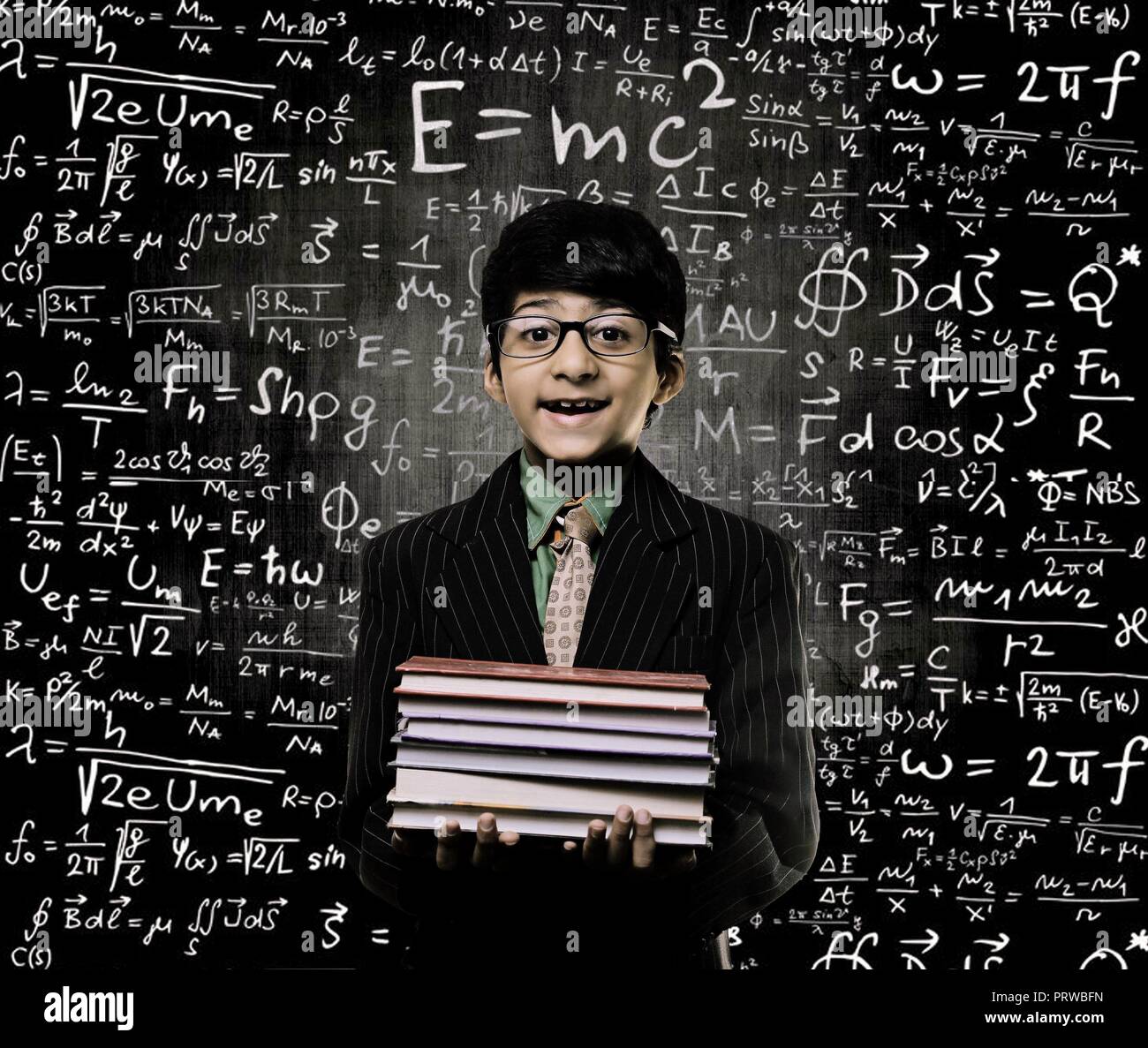 Cute Intelligent Little Boy Holding Book And Wearing Glasses Standing Before A Chalkboard, Chemical Formulas Are Written On Board Stock Photo