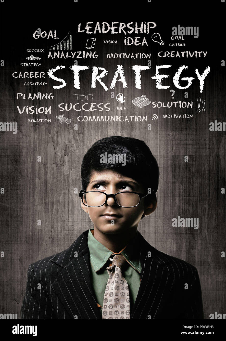 Cute Intelligent Little Boy Wearing Glasses On Nose, Thinking While Standing Before A Chalkboard, Word Strategy Written With Chalk Stock Photo