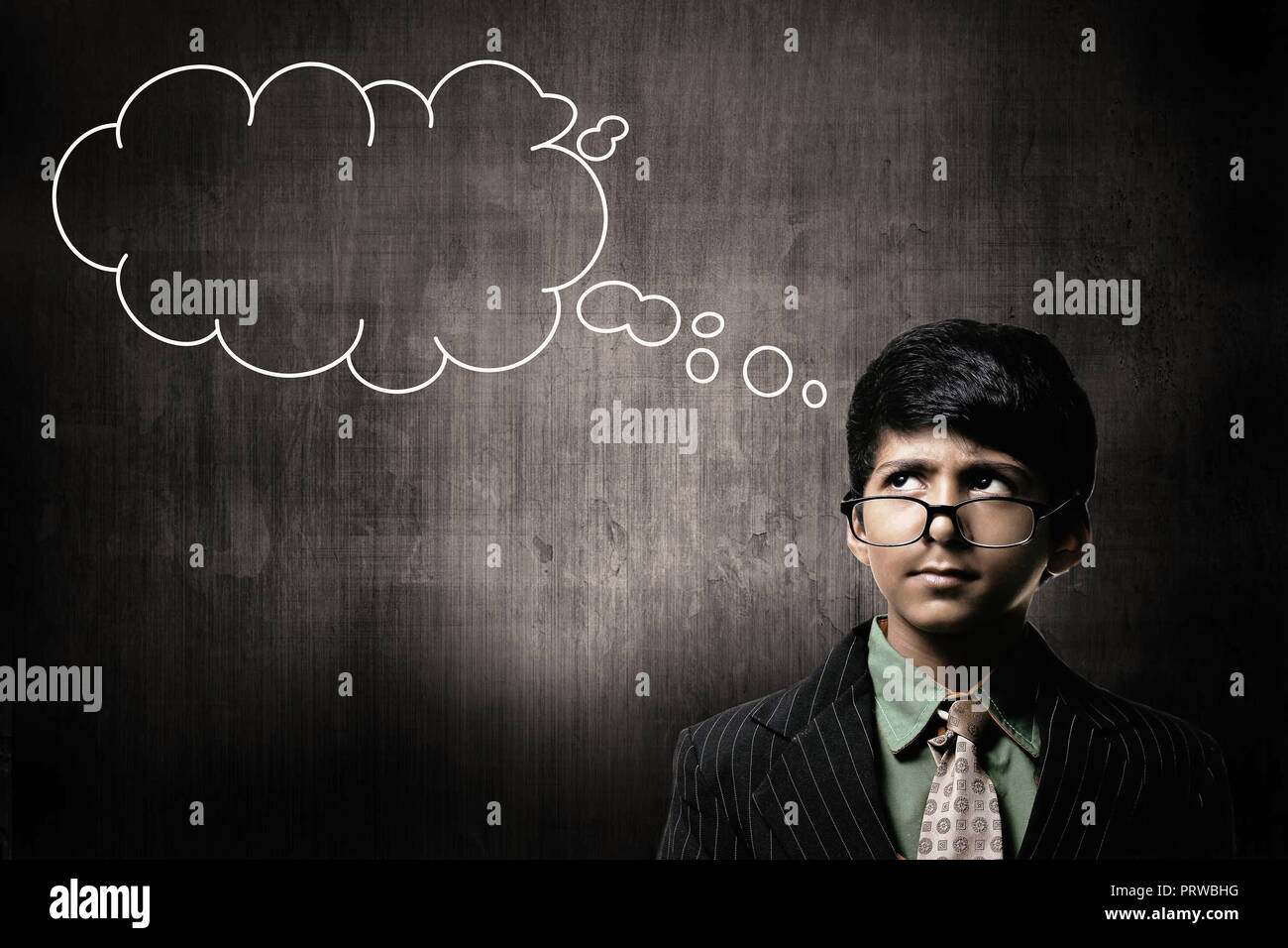 Cute Intelligent Little Boy Wearing Glasses On Nose, Thinking While Standing Before A Chalkboard, With Blank Thinking Cloud Stock Photo