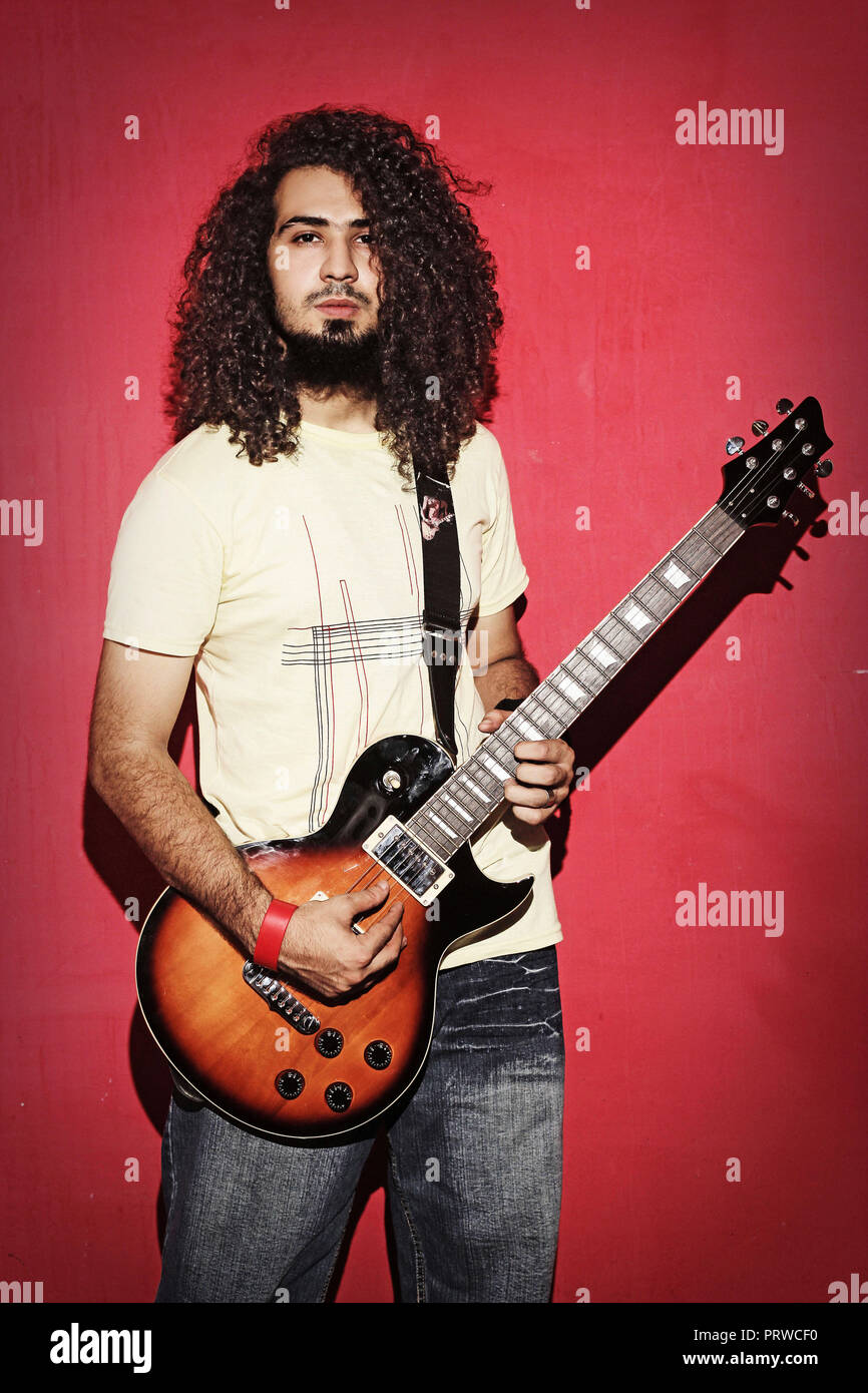 Closeup of one handsome passionate expressive cool young brunette  rock musician men with long curly hair playing electrIC guitar standing against red Stock Photo