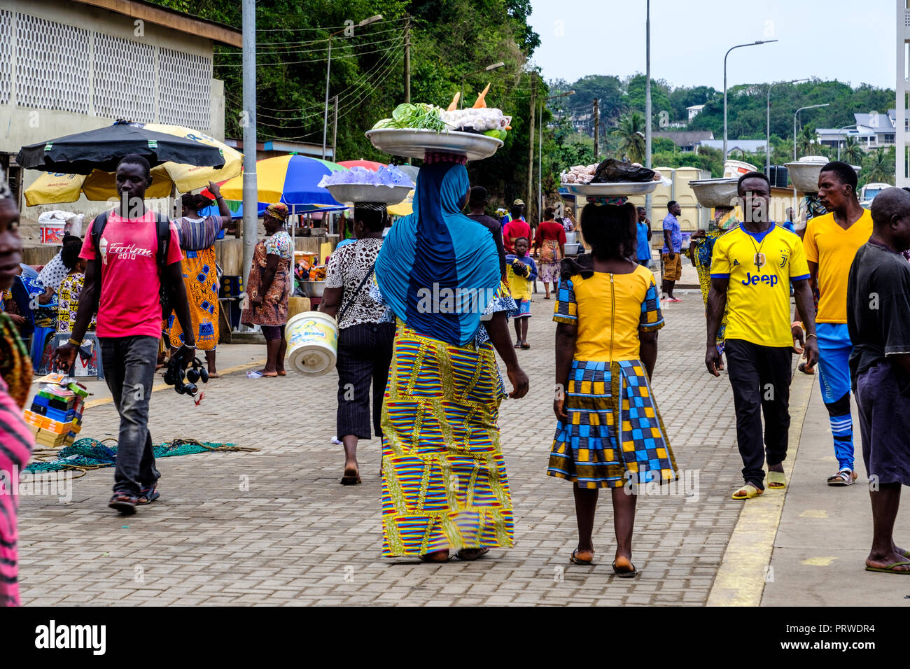 SEKONDI TAKORADI, GHANA – 10 APRIL 2018: Women in colorful dress carry baskets of produce on their heads ready to sell at busy Bosomtwi Sam Fishing Ha Stock Photo