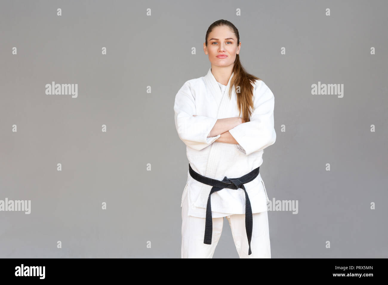 Beautiful confident athletic young woman in white kimono and black belt standing, crossed hands and looking at camera. Japanese martial arts concept.  Stock Photo