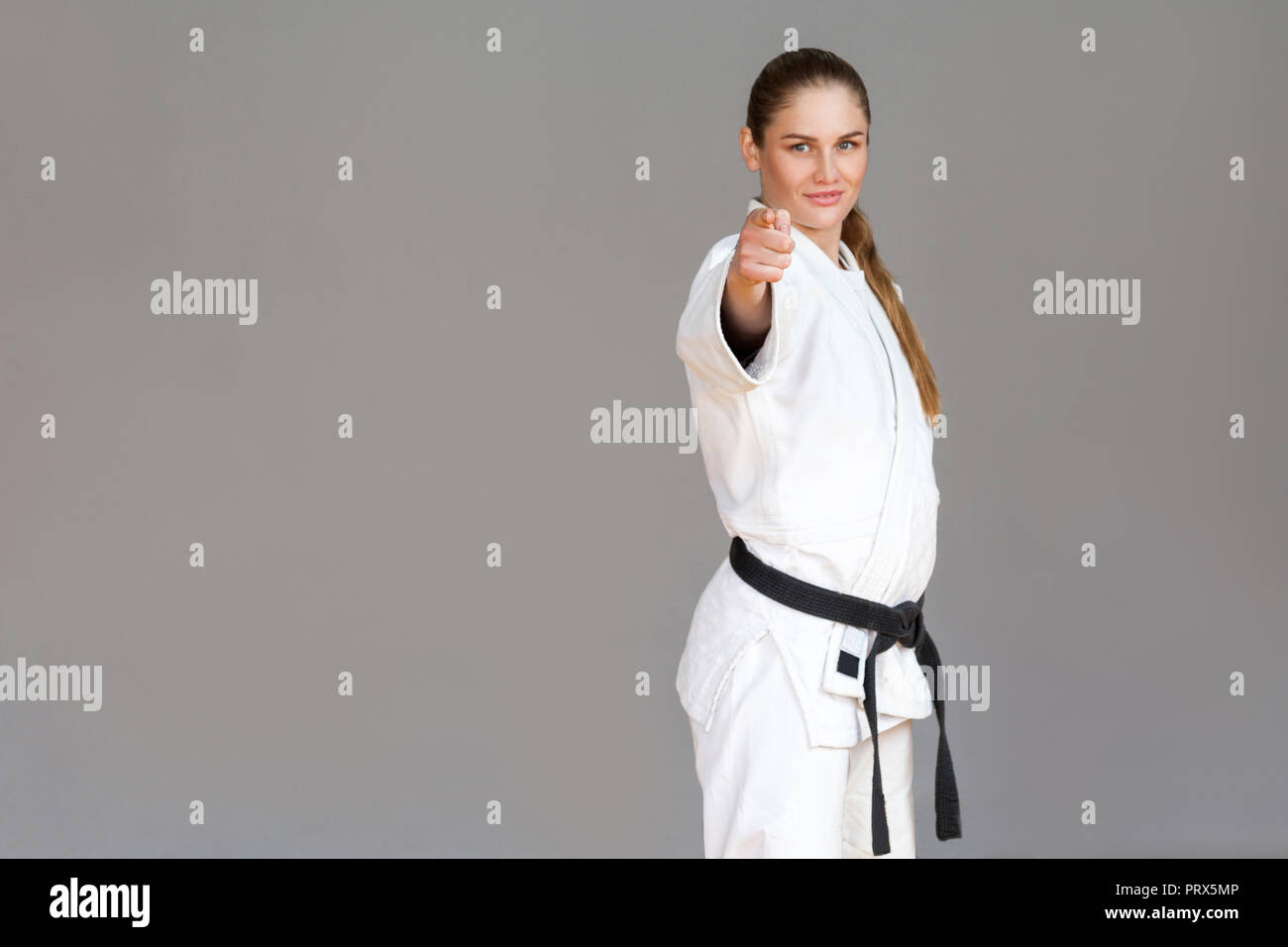 Confident athletic young woman in white kimono and black belt standing, pointing finger and looking at camera with smile. Japanese martial arts concep Stock Photo