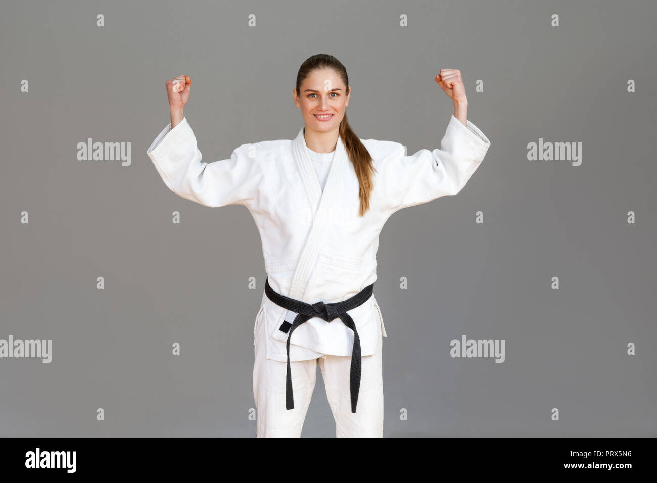 Happy confident athletic young woman in white kimono and black belt standing with raised arms showing strong hands looking at camera with toothy smile Stock Photo