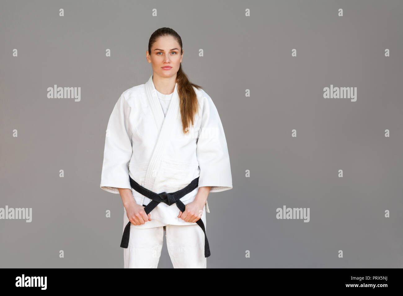 Beautiful athletic young woman in white kimono standing and holding black belt and looking at camera with serious face. Japanese martial arts concept. Stock Photo