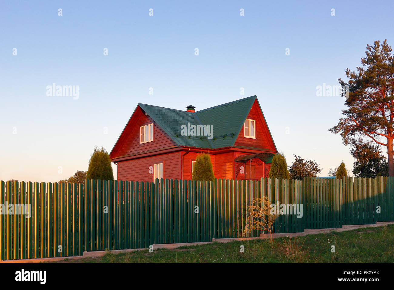 Wooden house under green metal roof house behind the green fence sunset lihgts photo Stock Photo