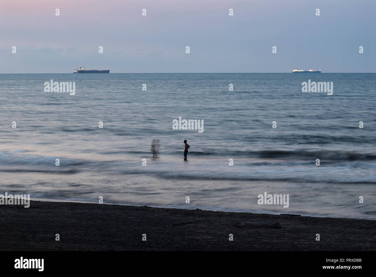 COATZACOALCOS, VER/MEXICO - SEPT 1st, 2018: Two man enjoy a dip at the beach during sunset. Oil tanker ships await entrance to the logistics terminal. Stock Photo