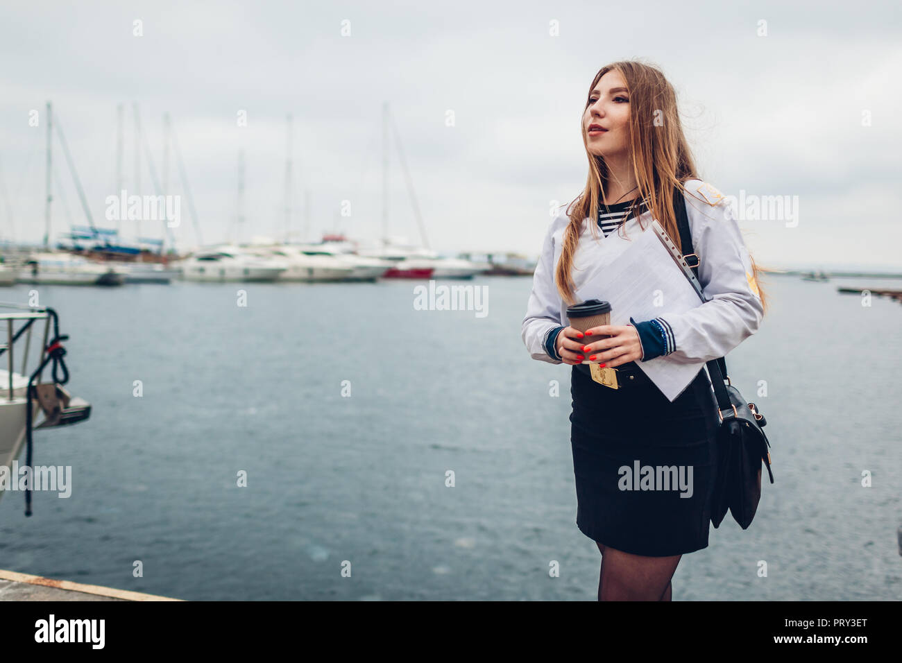 College woman student of Marine academy drinking coffee by sea wearing uniform. Girl walking in seaport of Odesa on pier Stock Photo