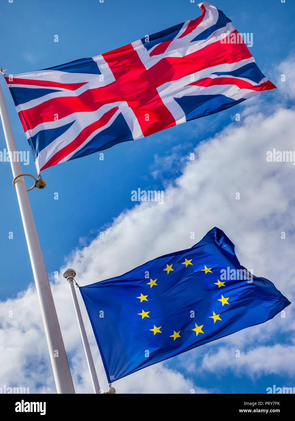 BREXIT FLAGS CONCEPT UK Union Jack Flag flying high above EU European Flag in a stiff breeze on a sunlit day with blue sky & flags divided by clouds Stock Photo