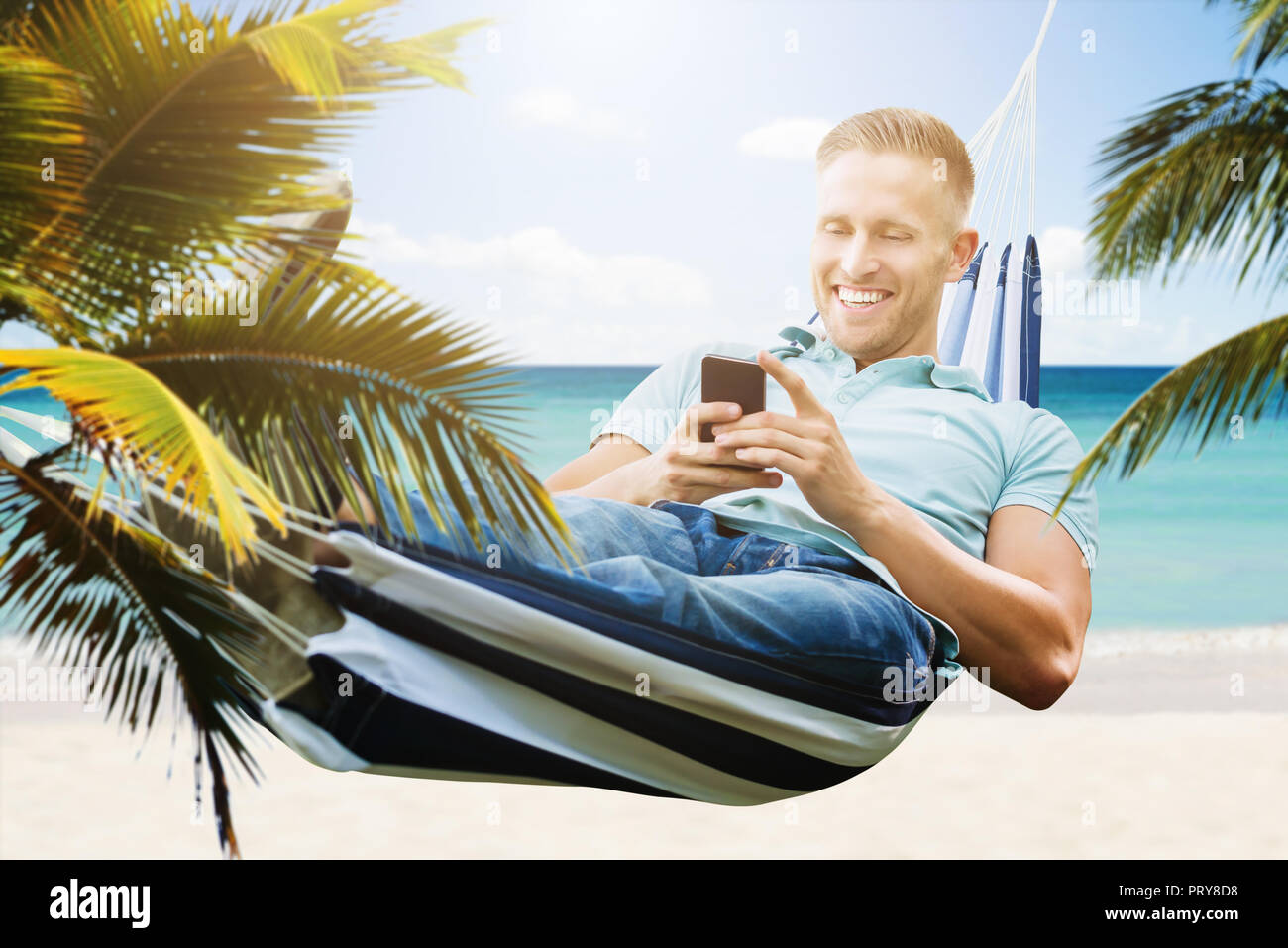 Happy Young Man Relaxing In Hammock Using Smartphone Stock Photo