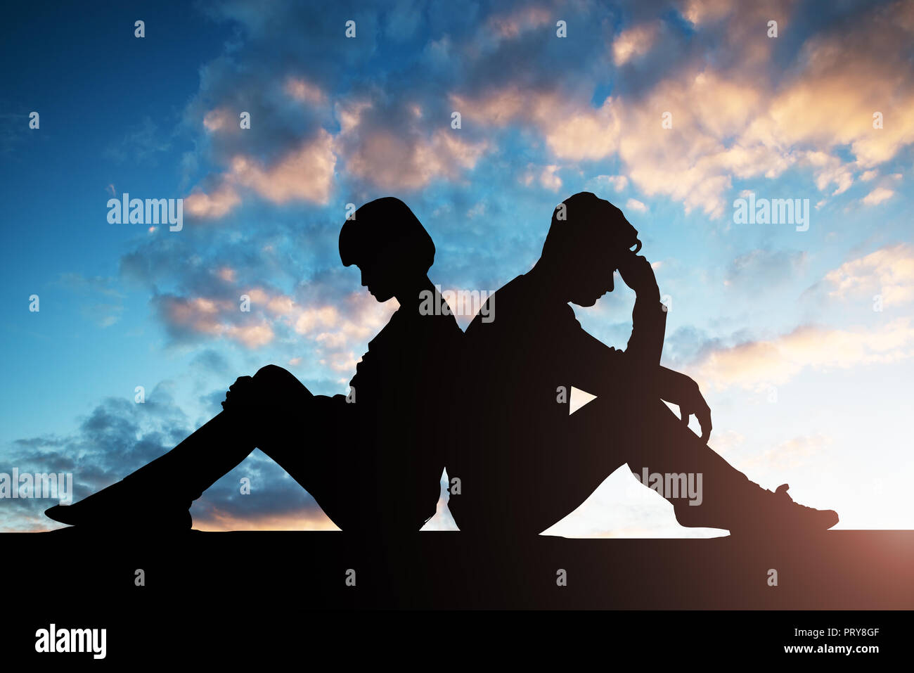 Silhouette Of Sad Couple Sitting Back To Back Against Cloudy Sky Stock Photo