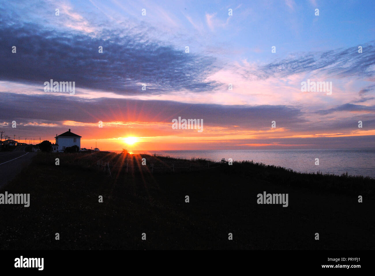 Blue, pink and orange cloudy sunset at the Gaspésie peninsula of Quebec, Canada, showing a black hill with a local house in contrast, and the calm sea Stock Photo