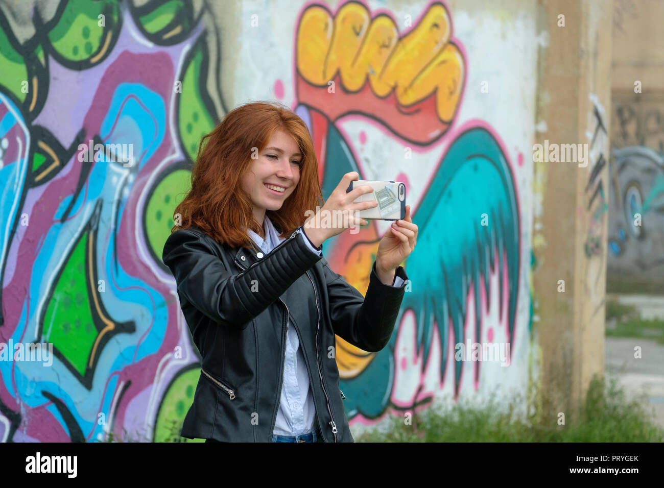Redheaded girl, young woman in leather jacket making a selfie with her smartphone in front of graffiti, near Finale Ligure Stock Photo