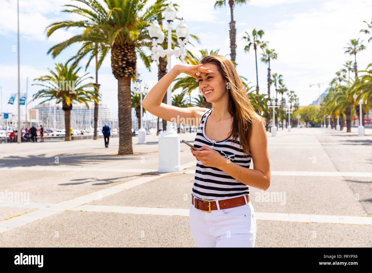 Smiling young woman holding smartphone at waterfront promenade Stock ...