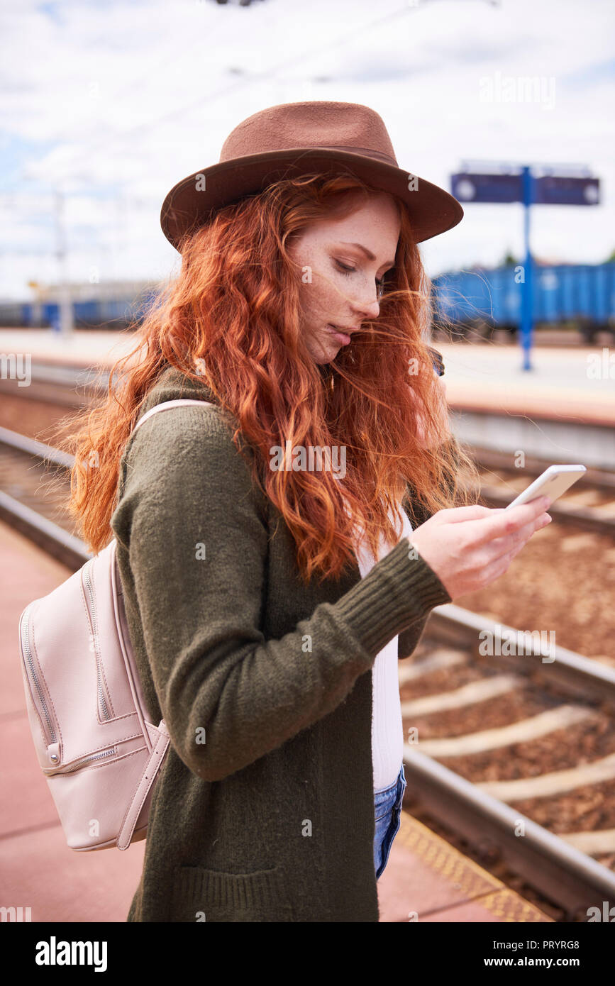 Redheaded woman wearing hat standing at platform looking at cell phone Stock Photo