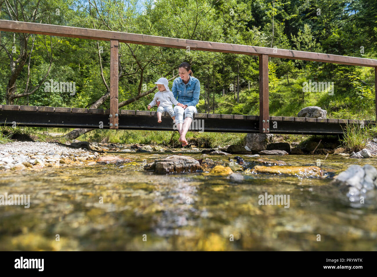 Mother and daughter sitting on wooden bridge, mountain stream Stock Photo