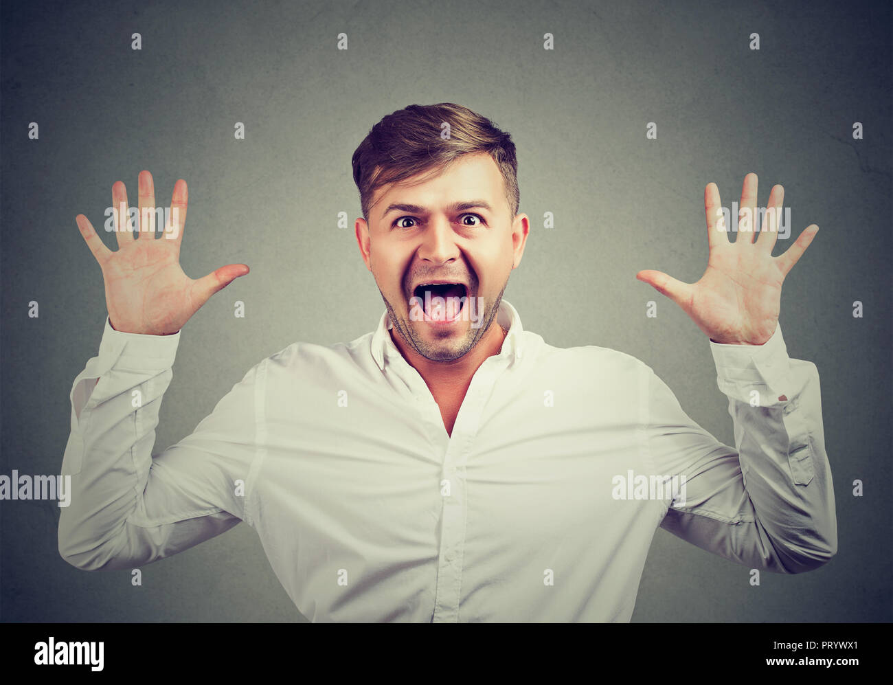 Young man in white shirt looking super impressed and surprised while screaming with hands raised at camera Stock Photo