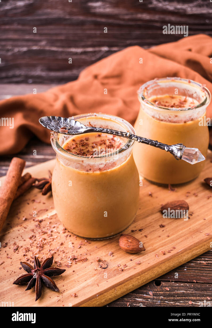 Toffee caramel dessert in jar, sprinkled with grated chocolate. On wooden board, decorated with cinnamon rolls, almonds and star anise. Rustic wooden  Stock Photo