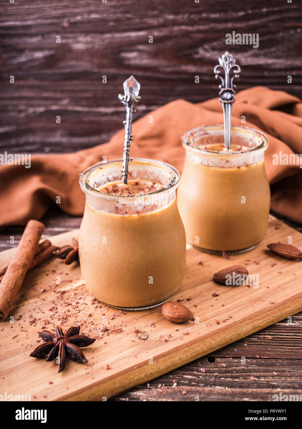 Toffee caramel dessert in jar, sprinkled with grated chocolate. On wooden board, decorated with cinnamon rolls, almonds and star anise. Rustic wooden  Stock Photo