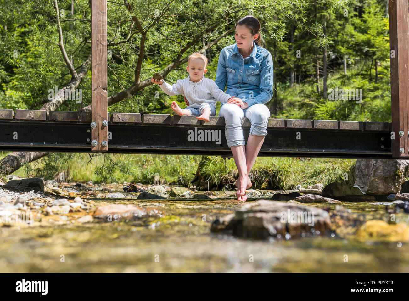 Mother and daughter sitting on wooden bridge, girl throwing stone into mountain stream Stock Photo