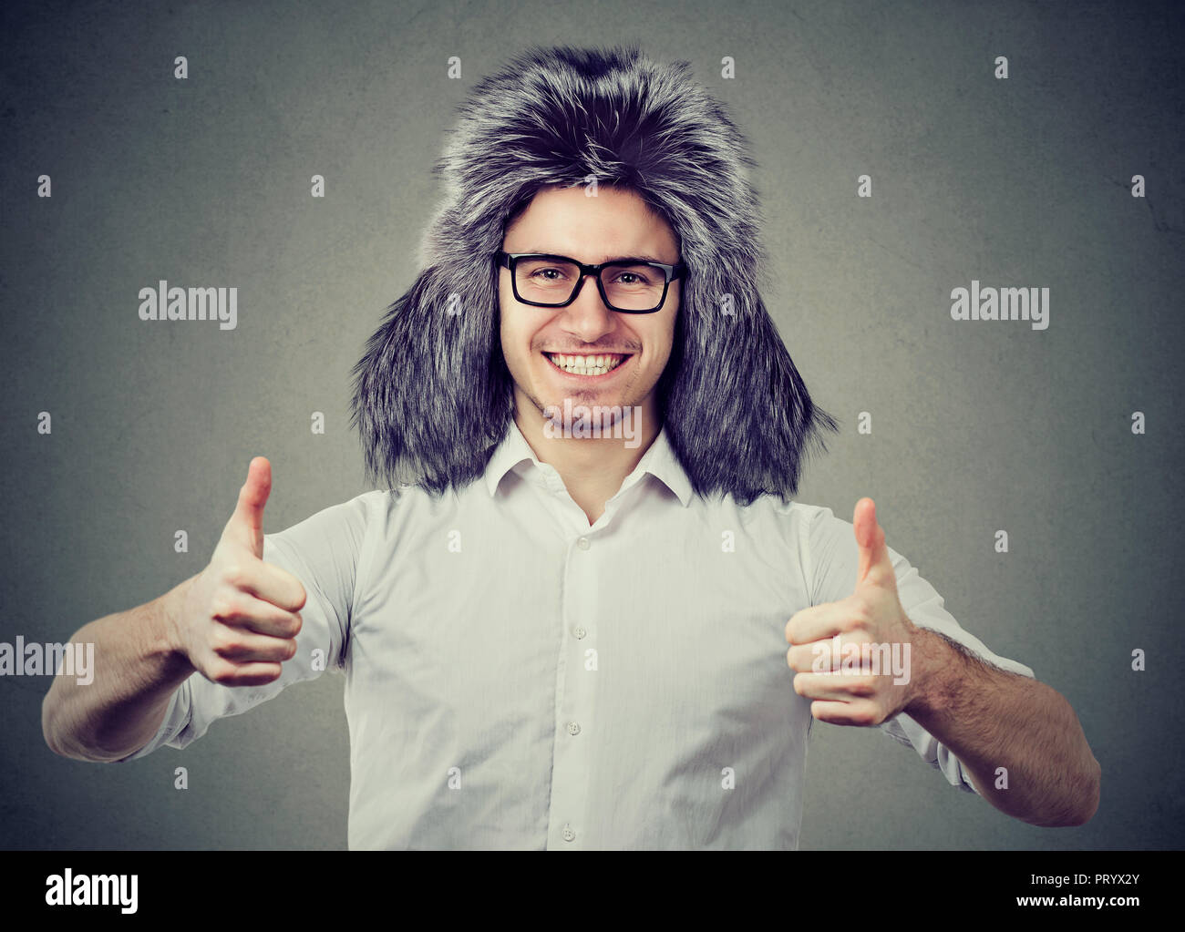 Young man wearing trendy furry winter hat and showing thumb up looking happily at camera on gray background Stock Photo