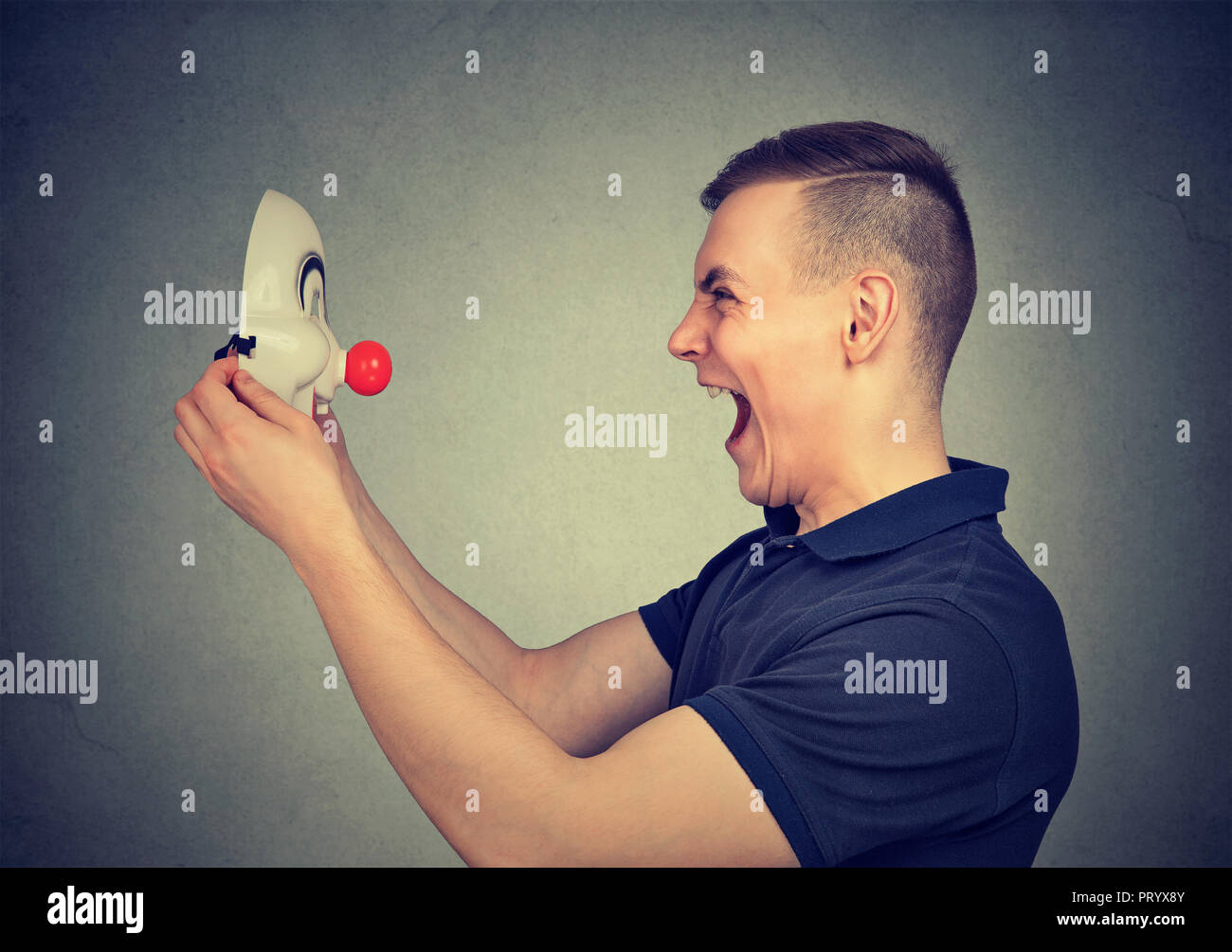 Side view of casual man yelling and looking at clown mask standing on gray background Stock Photo