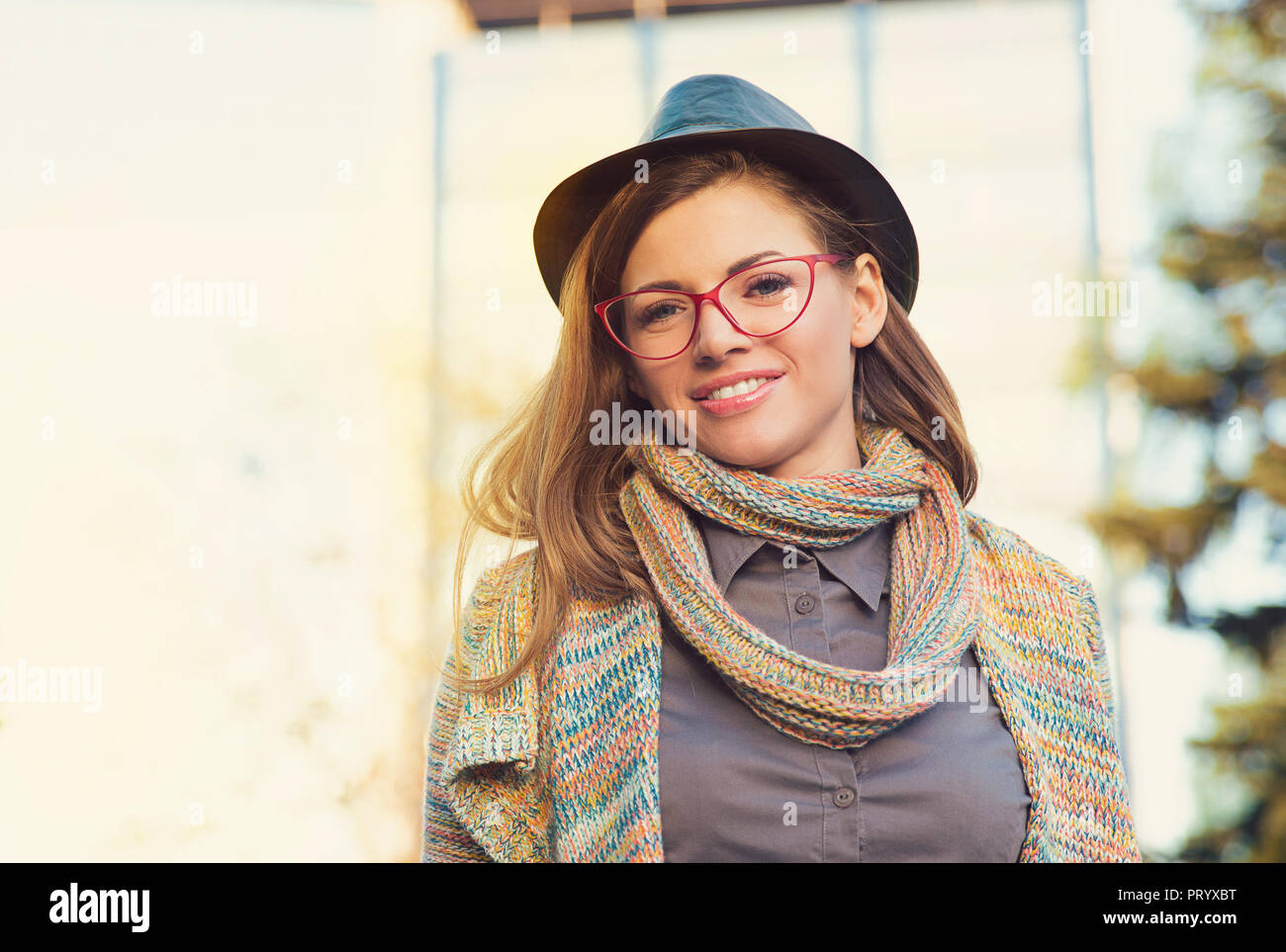 Charming modern woman wearing hat with glasses and scarf looking at camera in sunlight Stock Photo